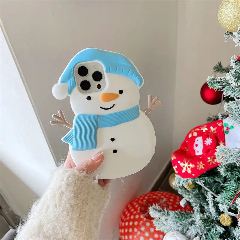 Phone case shaped like a snowman with a blue hat and scarf, held in front of a decorated Christmas tree.